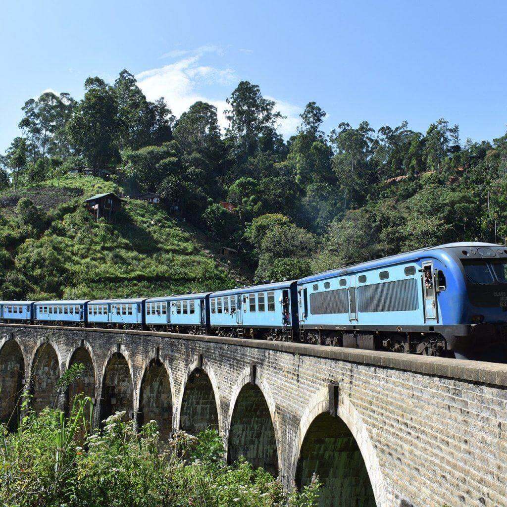 A blue train crossing a stone viaduct in the hills near Ella
