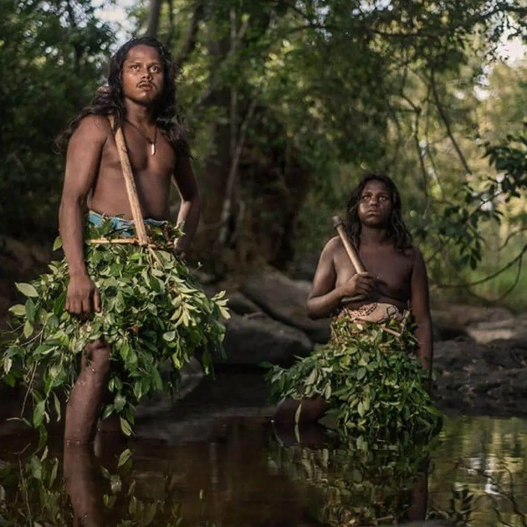 Two Vedda people standing in a stream in skirts of green leaves