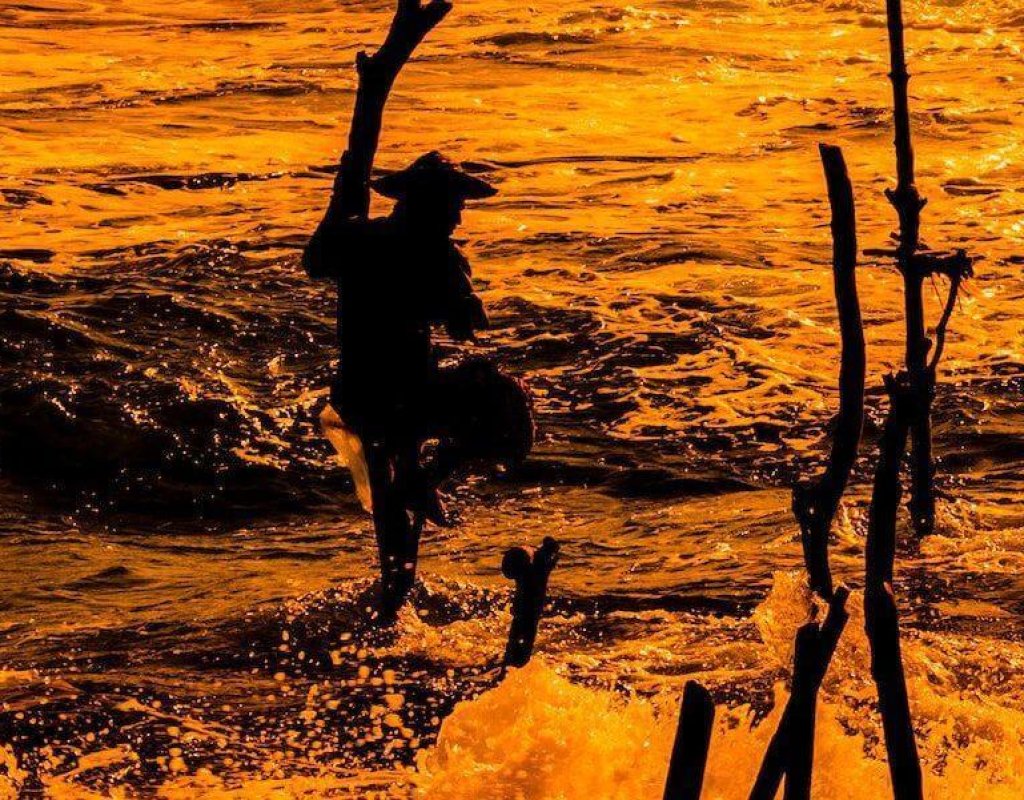 Silhouette of a stilt fisherman against orange sea spray at Mirissa