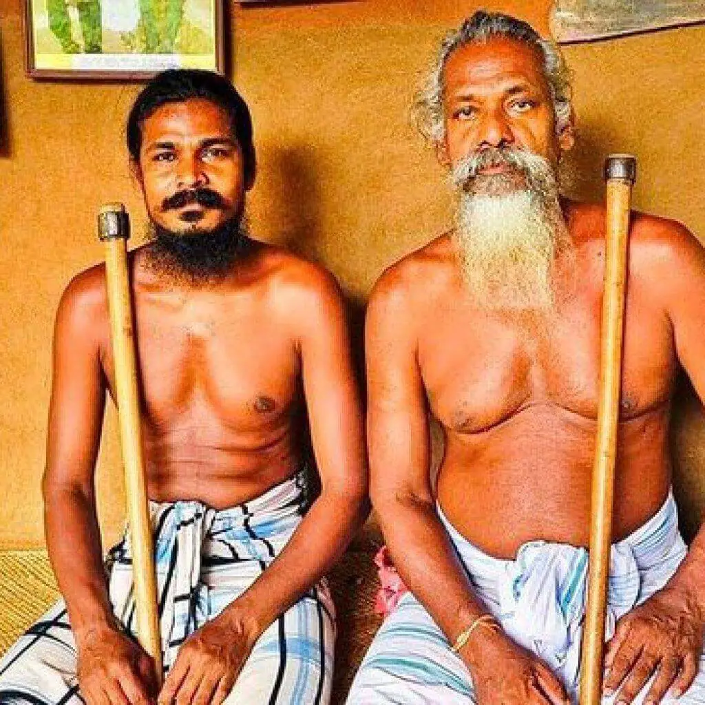 Two Vedda men sitting side by side holding walking staffs